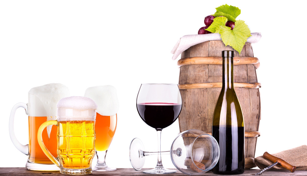 Weintrauben auf einem Weinfass, davor Gläser mit Bier und Wein sowie eine Flasche Rotwein mit Korkenzieher